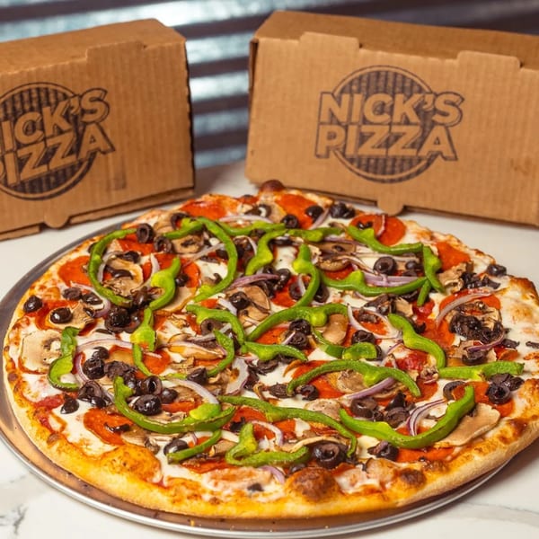 A Nick's Pizza pie with pepperoni, mushrooms, olives, onions, and green peppers, with two pizza boxes behind it.