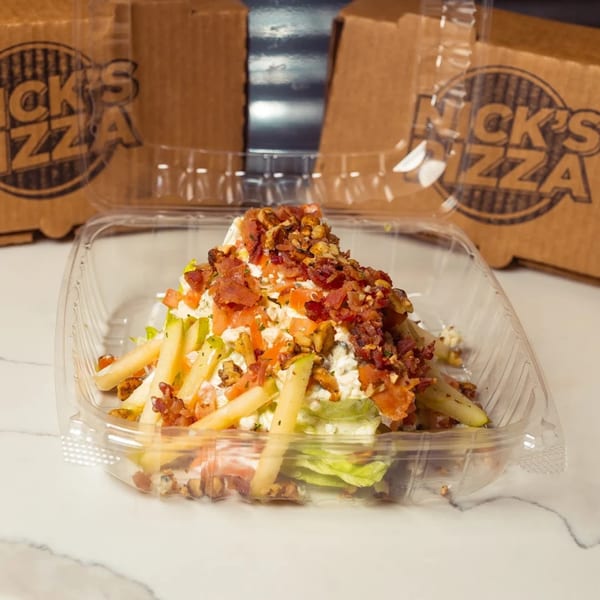 Nick's Pizza salad with bacon, apples, and cheese in a plastic container with pizza boxes behind it.